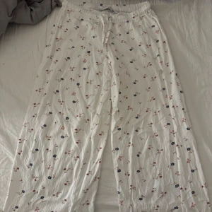 Vita blommiga byxor med snörning - Säljer ett par vita byxor med blommigt mönster i rosa och blått. Byxorna har en loose passform och är utrustade med snörning i midjan för justerbar passform. Perfekta för en avslappnad stil. Från lager 157.