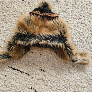 Pälsmössa från Canada Snow - Säljer en varm och fluffig pälsmössa från Canada Snow. Mössan är i svart och brun färg med stickade detaljer och en stor tofs på toppen. Perfekt för kalla vinterdagar.