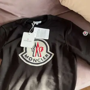 Säljer en snygg svart tröja från Moncler i nytt skick. Den har ett stort broderat Moncler-logotyp på framsidan i grått och rött. Tröjan är perfekt för både vardag och festliga tillfällen. Storleken är M och passar S 