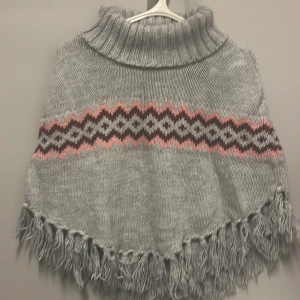 Grå stickad poncho med mönster - Säljer en supermysig grå stickad poncho med ett snyggt mönster i rosa och mörkrosa. Den har en hög polokrage och fransar längs nederkanten som ger en bohemisk touch. Perfekt för kyliga höst- och vinterdagar. Passar perfekt över en långärmad tröja för extra värme!