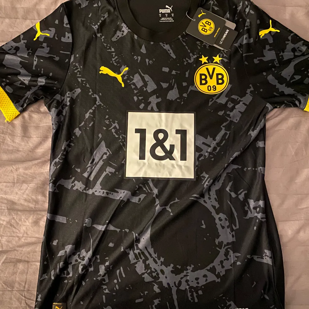 Säljer en svart Borussia Dortmund fotbollströja från Puma i nytt skick. Tröjan har korta ärmar och ett coolt mönster i grått. Den har klubbens emblem och sponsorlogga på framsidan, samt 'Dortmund' och 'Sancho 10' på baksidan. Perfekt för alla BVB-fans! 🖤💛. Sportswear.