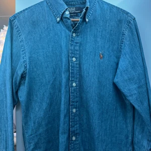 Blå jeansskjorta från Polo Ralph Lauren - Säljer en snygg blå jeansskjorta från Polo Ralph Lauren i storlek M. Skjortan har en slim fit passform och är långärmad med klassiska knappar framtill. Perfekt för både vardag och fest! 💙