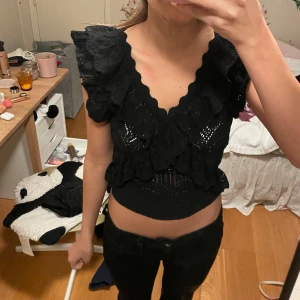 Svart volangtopp - Säljer en snygg svart topp med volanger och djup v-ringning. Toppen är tight och har ett fint mönster som ger en elegant look. Perfekt för festliga tillfällen!