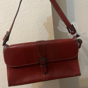 Röd axelväska i skinn - Säljer en snygg vinröd axelväska med justerbar rem och spänne framtill. Väskan har en stilren design med vita sömmar som ger en fin kontrast. Perfekt för både vardag och fest! Lite slitage på remmen, men annars i bra skick.