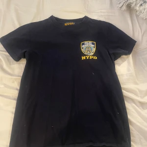 Svart NYPD t-shirt - Säljer en svart NYPD t-shirt i 100% bomull. Den har en officiell licensierad logga på bröstet och texten 'NYPD' i gult. Perfekt för en avslappnad stil eller som en cool present. Passar bra till jeans eller shorts!