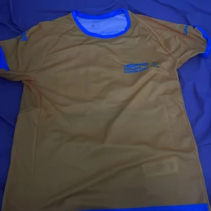 Gul t-shirt från Korpen - Säljer en gul t-shirt från Korpen med blå detaljer och texten 'Motionslandslaget'. Den har nummer 4109 tryckt på baksidan. Perfekt för träning eller löpning. T-shirten är kortärmad och i bra skick.