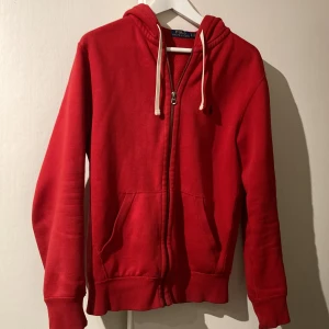 Röd hoodie från Polo Ralph Lauren - Säljer en snygg röd hoodie från Polo Ralph Lauren i ganska bra skick. Den har en dragkedja framtill och en klassisk logga broderad på bröstet. Perfekt för höst och vår, med långa ärmar och en skön passform. Huvan har vita snören för justering. Passar både till vardags och chill. Om den är för lite åker dragkedjan ner lite när man har luvan på