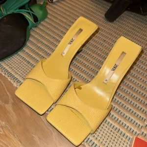 Gula klackskor från Zara - Snygga gula klackskor från Zara i storlek 38. De har en fyrkantig tå och en stilren design som passar perfekt för sommarens fester. Skorna är välanvända och ena sulan har släppt men går att limma tillbaka. Perfekta för att liva upp vilken outfit som helst!