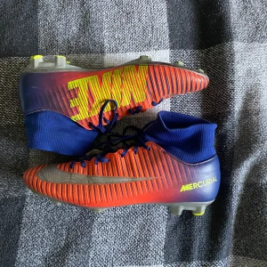 Nike Mercurial fotbollsskor - Säljer ett par Nike Mercurial fotbollsskor i rött och blått med gula detaljer. Skorna har en tight passform och snörning för bästa komfort på planen. De är i bra skick med lite slitage på sulorna. Perfekta för fotbollsträning eller match!