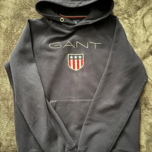 S Svart hoodie från GANT - Säljer en snygg svart hoodie från GANT med deras ikoniska logga och ett sköldmärke på bröstet. Den är i bra skick och perfekt för höst och vinter. Hoodien har en klassisk passform med långa ärmar och en bekväm huva. Perfekt för både vardag och chill! 🖤