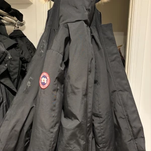 Svart parkajacka med pälsdetalj - Säljer en svart parkajacka från Canada Goose i mycket bra skick. Jackan har en stor huva med pälsdetalj och är perfekt för kalla vinterdagar. Den har en dragkedja framtill och är både stilren och funktionell. Passar perfekt för vinterns alla äventyr! Skick 9/10 