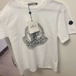 Säljer en snygg vit t-shirt från Moncler i nytt skick. Den har ett stort Moncler-logotryck på framsidan och en liten patch på ärmen. Perfekt för en stilren och avslappnad look. Passar till både vardag och fest!