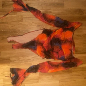 Färgglad body med knytband - En body i härliga färger som orange, röd och svart. Den har långa ärmar och ett coolt mönster som verkligen sticker ut. Perfekt för en fest eller en utekväll! 