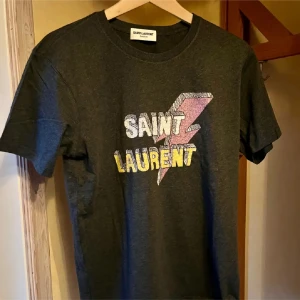 t-shirt från Saint Laurent storlek M - Säljer en grå t-shirt från Saint Laurent med ett coolt blixttryck och texten 'SAINT LAURENT' i vitt och gult. T-shirten är i nytt skick och perfekt för en avslappnad stil. Tillverkad i Italien och har en normal passform. Perfekt för både vardag och fest!