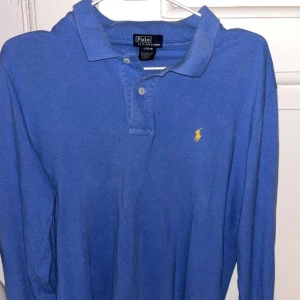 Blå pikétröja från Ralph Lauren - Säljer en klassisk blå pikétröja från Ralph Lauren i barnstorlek L men passar S i herr Tröjan är i bomull och har långa ärmar med Ralph lauren loggan på bröstet. Perfekt för både vardag och lite finare tillfällen. Skön och stilren design!