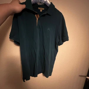 Blå piké från burberry - Säljer en snygg pikétröja i bra skick. Den har en klassisk krage med det ikoniska burberry mönstret och korta ärmar, perfekt för både vardag och lite finare tillfällen.  Passar perfekt till jeans eller chinos! Skickar gärna fler bilder om det skulle önskas!