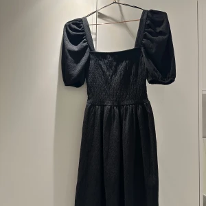 Svart klänning från H&M - Snygg svart klänning från H&M i storlek XS. Den har puffiga kortärmar och en smickrande passform med rynkad detalj över bysten. Perfekt för både vardag och festliga tillfällen. Materialet känns mjukt och bekvämt mot huden.