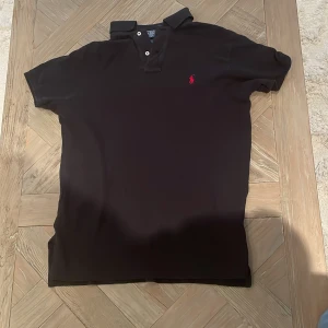 Svart pikétröja från Polo Ralph Lauren - Säljer en klassisk svart pikétröja från Polo Ralph Lauren i storlek M. Tröjan har en röd broderad logga på bröstet och knappar vid kragen. Perfekt för en stilren och avslappnad look. Passar både till vardags och fest!