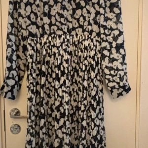 Svartvit blommig klänning - Säljer en elegant svartvit klänning med blommigt mönster. Klänningen har långa ärmar och ett plisserat kjolparti som ger en fin rörelse. Perfekt för både vardag och festliga tillfällen. Materialet känns mjukt och bekvämt mot huden. Fint skick förutom att den har tappat en knapp längst ner på vänster arm, det går lätt sy på en ny knapp och inget som man stör sig på. 