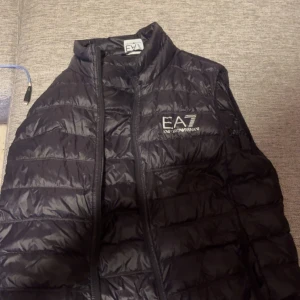 Svart dunväst från Emporio Armani - Snygg svart dunväst från Emporio Armani med EA7-logga på bröstet. Västen har en quiltad design och dragkedja framtill. Perfekt för att hålla värmen med stil.