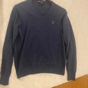 Blå tröja från Gant - Säljer en stilren blå tröja från Gant med v-ringad hals. Tröjan har långa ärmar och ribbade muddar vid ärmslut och nederkant. Perfekt för en avslappnad men snygg look.