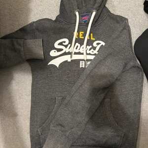 Grå hoodie från SuperDry - Säljer en snygg grå hoodie från SuperDry med vit och gul text på framsidan. Tröjan har en klassisk känguruficka och justerbar huva med snören. Perfekt för en avslappnad stil. Väldigt unik och exklusiv och kan definitivt diskutera pris✅✅✅✅✅🔥🔥🔥🔥🔥🔥🔥❤️❤️❤️❤️❤️