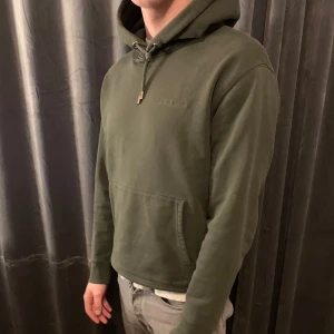 Olivgrön hoodie från J. Lindeberg - Säljer en stilren grön hoodie från J.Lindeberg. Den har en klassisk design med justerbar huva med snörning. Perfekt för en avslappnad look.