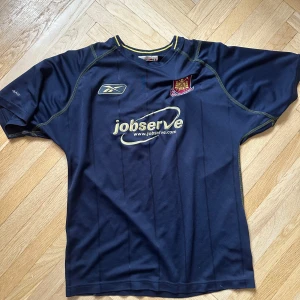 Mörkblå fotbollströja från Reebok - Säljer en mörkblå fotbollströja från Reebok med korta ärmar och klubbmärke på bröstet. Tröjan har gula sömmar och reklamtryck på framsidan. 