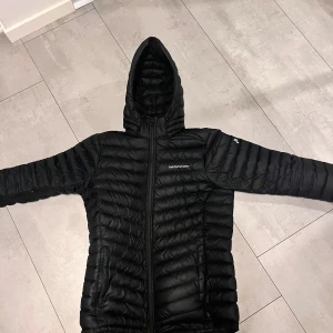 Svart dunjacka från Peak Performance - Säljer en stilren svart dunjacka från Peak Performance. Jackan har en praktisk huva och dragkedja framtill. Perfekt för kyliga dagar med sin lätta och värmande design. Passar både till vardags och utomhusaktiviteter.