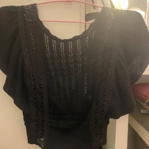 Svart spetsblus - Svart blus med spetsdetaljer och korta ärmar. Öppen i ryggen. Använd endast en gång, väldigt bra skick. 