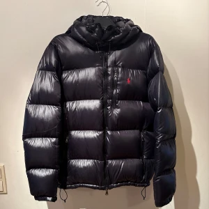 Svart dunjacka från Ralph Lauren - Säljer en snygg svart dunjacka från Ralph Lauren med en röd broderad logga på bröstet. Jackan är i storlek L, skick 8/10 då den har en liten öppning på armen men som man ser på bilden har jag lagat den och man ser knappt att den har gått sönder! Hör av dig vid flera frågor✅