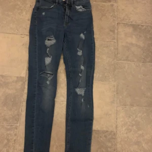 Blå jeans från H&M - Snygga blå jeans från h&m med slitna detaljer längs benen. De har en klassisk femficksdesign och knappgylf. Perfekta för en avslappnad stil.