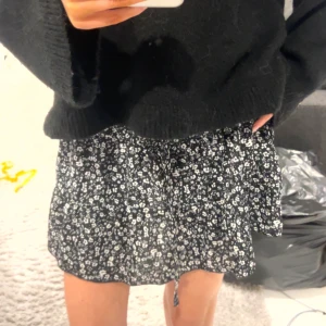 Svart blommig kortkjol - Säljer en söt svart kortkjol med vita blommor. Kjolen har en avslappnad passform och är perfekt för en trendig look. Den är lätt och luftig, vilket gör den idealisk för varmare dagar.