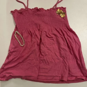 Rosa ärmlös topp med smockdetaljer - Söt rosa ärmlös topp med smockdetaljer upptill och tunna axelband. Toppen har en dekorativ guldfärgad blomma på bröstet. Perfekt för varma sommardagar!