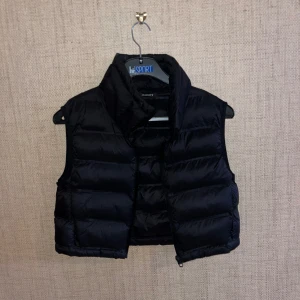 Svart dunväst från Zara - Snygg svart dunväst från Zara med en croppad design. Perfekt för lager-på-lager under kyligare dagar. Västen har en hög krage och stängs med dragkedja framtill.