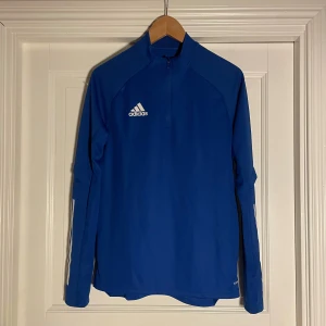 Blå långärmad tröja från Adidas - Säljer en blå långärmad tröja från Adidas med dragkedja och logotyp på bröstet. Tröjan är tillverkad i syntetmaterial och har en sportig design, perfekt för träning eller vardagsbruk. Adidas AEROREADY-teknologi för optimal komfort.