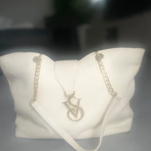 Vit axelväska med kedjedetaljer - Snygg vit axelväska med guldfärgade kedjedetaljer och ett stort VS-emblem framtill. Väskan har en praktisk dragkedja inuti och gott om plats för dina saker. Perfekt för att ge din outfit en stilren touch.