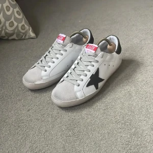 Vita sneakers med svart stjärna - Snygga vita sneakers från Golden Goose med en svart stjärna på sidan och grå snörning. Skorna har en svart häl och en diskret logga på sidan. Perfekta för en stilren look.