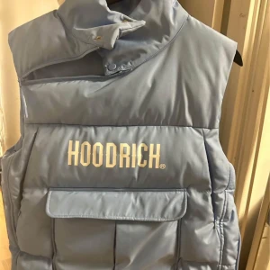 Ljusblå dunväst från Hoodrich - Snygg ljusblå dunväst från Hoodrich med hög krage och tryckknappar. Västen har en stor ficka framtill och är perfekt för kyligare dagar. Den har en modern och stilren design.