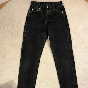 Svarta Levis 501 jeans - Snygga svarta Levis 501 jeans med klassisk design och knappgylf.  De har en hög midja och raka ben.                                                          W 28 L 32  (Dock nedkortade till 31)   Jeansen är i gott skick. Enda anmärkningarna är att dem är uppsydda 1 tum och att det har gjorts en liten slits längst ner i byxan. (Se sista bilden)   Längd hela byxan: 103 cm Längd ben innersöm: 78 cm  Vidd runt midjan: 71 cm  100% bomull