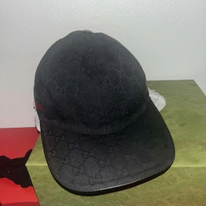 Svart Gucci keps - Snygg svart keps från Gucci med diskret mönster och klassiska gröna och röda ränder på sidan. Perfekt för att ge en stilren touch till din outfit. Kepsen har en rundad skärm och justerbar passform. 
