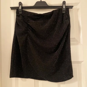 Svart glittrig kjol från Kappahl - Snygg svart glittrig kjol från Kappahl i en kort modell. Perfekt för att lysa upp vilken outfit som helst. Kjolen har en enkel design med en glittrig yta som ger en festlig känsla. Ett måste för garderoben!