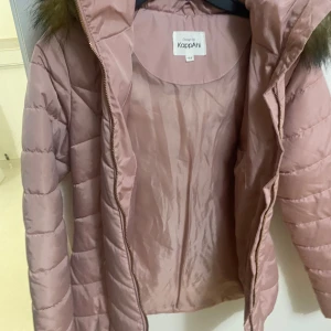 Rosa quiltad jacka från KappAhl - Säljer en snygg rosa quiltad jacka från KappAhl med en mysig pälskantad huva. Jackan har dragkedja framtill och är perfekt för kyligare dagar. Den är både stilren och praktisk med sin varma design.