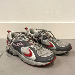 Grå och röda sneakers från Saucony - Snygga och sportiga sneakers från Saucony i grått och rött. Skorna har en robust design med snörning och en bekväm passform. Perfekta för en aktiv livsstil med bra grepp och stöd.