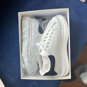 Vita sneakers från Alexander McQueen - Snygga vita sneakers från Alexander McQueen med svart detalj på hälen. Skorna har en klassisk design med snörning och en tjock sula. Perfekta för att ge en stilren touch till din outfit.