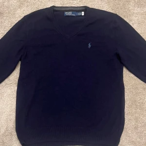 Mörkblå v-ringad tröja från Ralph Lauren - Säljer en stilren mörkblå tröja från Ralph Lauren med v-ringning och broderad logga på bröstet. Tröjan har långa ärmar och är perfekt för en klassisk look. Passar bra till både jeans och chinos samt skjorta under. Nypris ~ 2000kr, storlek M, 100% ull. 