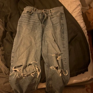 Blå slitna jeans - Säljer ett par blå jeans med slitningar på knäna och låren. De har en klassisk femficksdesign och en knappgylf. Perfekta för en avslappnad stil.