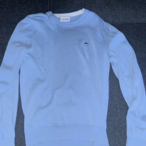 Ljusblå tröja från Lacoste - Säljer en stilren ljusblå tröja från Lacoste med rund halsringning och det klassiska krokodilmärket på bröstet. Tröjan har långa ärmar och ribbade muddar vid ärmslut och nederkant. Perfekt för en avslappnad och snygg look. Köpt för 1200kr