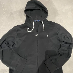 Svart hoodie från Polo Ralph Lauren - Säljer en stilren svart hoodie från Polo Ralph Lauren. Varsamt använd och säljer nu då den aldrig kommer till användning. Skicket är mycket bra - men den är lite dammig, vilket jag givetvis tar bort vid köp. Hör av dig vid intresse eller ställ en fråga!