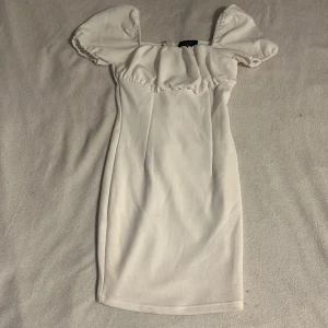 Vit klänning från AX Paris - Säljer en söt vit klänning från AX Paris med puffiga kortärmar och en dragkedja i ryggen. Klänningen har en tight passform och är perfekt för en stilren look. Den är tillverkad i ett mjukt material som ger en bekväm känsla. Endast envänd en gång på skolavslutningen förra året sen dess har den bara hängt i garderoben